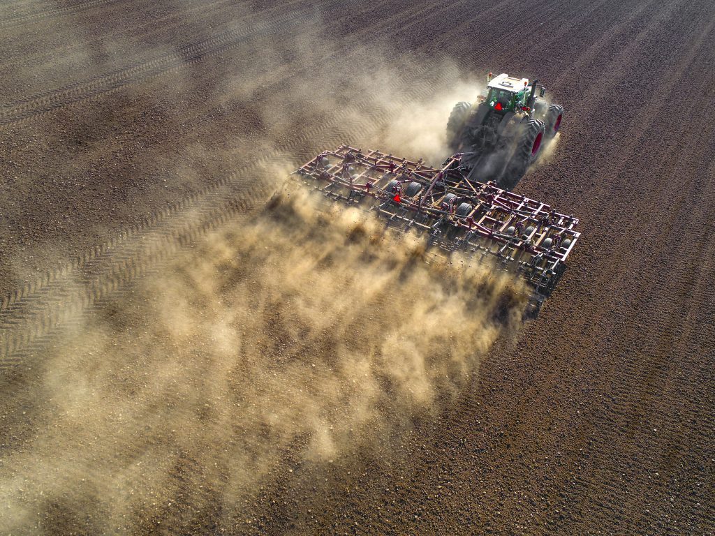 Figure 1: Big farm tractor tilling dusty Springtime fields, aerial view.